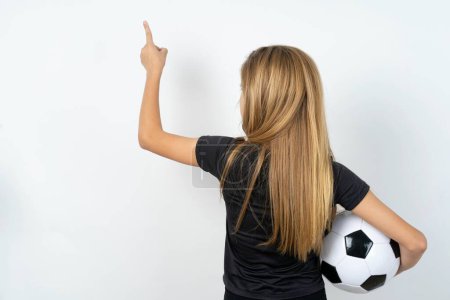 Téléchargez les photos : Adolescente portant des vêtements de sport tenant un ballon de football sur un mur blanc pointant vers l'objet sur l'espace de copie, vue arrière. Tourne le dos - en image libre de droit