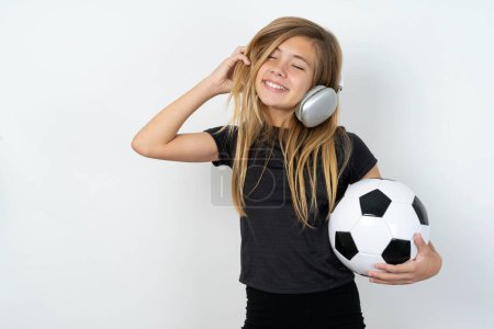 Téléchargez les photos : Adolescent fille portant des vêtements de sport tenant un ballon de football sur le mur blanc sourit largement se sent très heureux écoute piste de musique préférée via casque sans fil ferme les yeux. - en image libre de droit