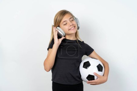 Photo for Teen girl wearing sportswear holding a football ball over white wall with headphones on her head, listens to music, enjoying favourite song with closed eyes, holding hands on headset. - Royalty Free Image
