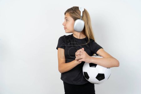 Téléchargez les photos : Adolescente portant des vêtements de sport tenant un ballon de football sur un mur blanc porte un casque stéréo écoutant de la musique concentrée et regardant de côté avec intérêt. - en image libre de droit