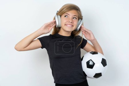 Téléchargez les photos : Joyeuse adolescente portant des vêtements de sport tenant un ballon de football sur un mur blanc chante chanson garde la main près de la bouche comme si le microphone écoute la playlist préférée via un casque - en image libre de droit