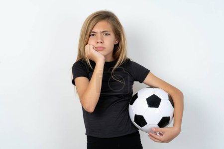 Foto de Retrato de una triste adolescente con ropa deportiva sosteniendo una pelota de fútbol sobre la cara de las manos blancas de la pared - Imagen libre de derechos