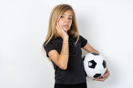 Téléchargez les photos : Photo de jeune fille portant des vêtements de sport tenant un ballon de football sur un mur blanc profiter d'une peau fraîche et lisse toucher visage - en image libre de droit