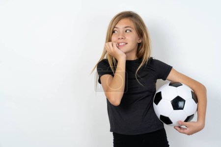 Photo for Terrified teen girl wearing sportswear holding a football ball over white wall looks empty space home alone moonless night - Royalty Free Image