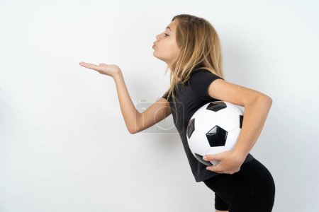 Téléchargez les photos : Profil vue de côté, portrait d'une jolie adolescente portant des vêtements de sport tenant un ballon de football sur un mur blanc envoyant un baiser d'air - en image libre de droit