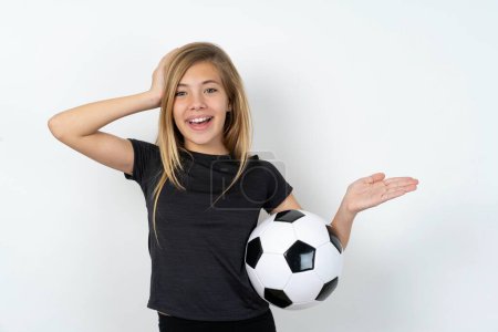 Foto de Sorprendido sorprendido adolescente sorprendido usando ropa deportiva sosteniendo una pelota de fútbol sobre la pared blanca celebrar la mano ofreciendo proposición - Imagen libre de derechos
