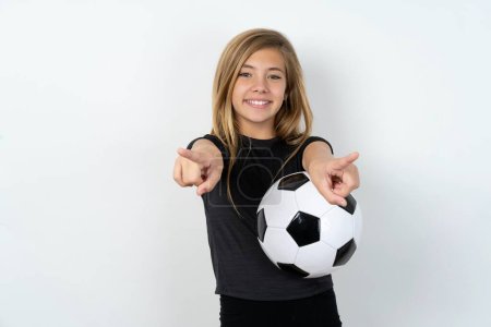Téléchargez les photos : Adolescent fille portant des vêtements de sport tenant un ballon de football sur blanc mur joyeux et souriant pointant vers la caméra - en image libre de droit