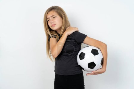 Foto de Adolescente chica usando ropa deportiva celebración de un balón de fútbol sobre blanco pared tiene dolor de espalda - Imagen libre de derechos