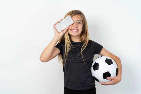 Photo for Teen girl wearing sportswear holding a football ball over white wall holding modern smartphone covering one eye while smiling - Royalty Free Image