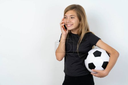 Photo for Happy joyful charming teen girl wearing sportswear holding a football ball over white wall speak phone smile good mood - Royalty Free Image