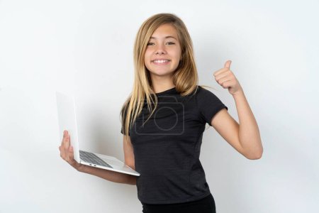 Téléchargez les photos : Belle caucasien adolescent fille portant noir t-shirt tenant netbook et montrant pouce vers le haut - en image libre de droit