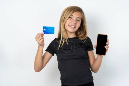Foto de Chica adolescente caucásica con camiseta negra abrió cuenta bancaria, la celebración de teléfono inteligente y tarjeta de crédito, sonriendo, recomiendan el uso de la aplicación de compras en línea en el estudio - Imagen libre de derechos