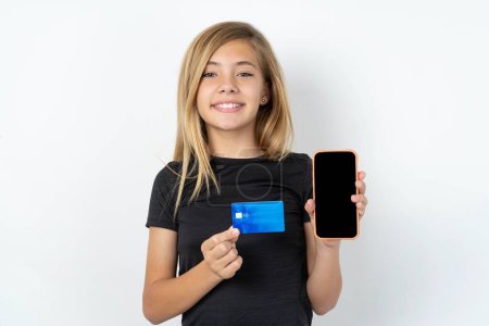Téléchargez les photos : Photo d'adorable adolescente caucasienne portant un T-shirt noir sur un mur blanc tenant une carte de crédit et un smartphone. Réservé aux achats en ligne - en image libre de droit