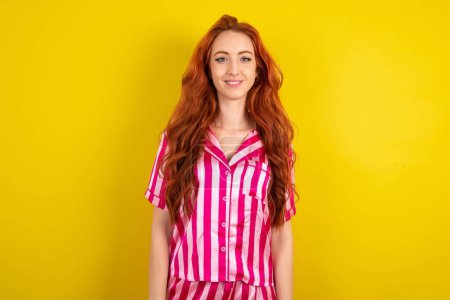 Téléchargez les photos : Jeune femme aux cheveux rouges portant un pyjama rose sur fond de studio jaune avec des dents souriantes - en image libre de droit