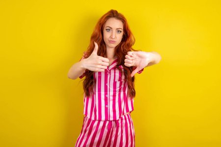 Téléchargez les photos : Jeune femme aux cheveux rouges portant un pyjama rose sur fond jaune studio montrant les pouces vers le haut et les pouces vers le bas, concept de choix difficile - en image libre de droit