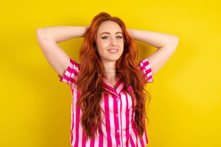Téléchargez les photos : Jeune femme aux cheveux rouges portant un pyjama rose sur fond jaune studio étirant les bras, position détendue. - en image libre de droit