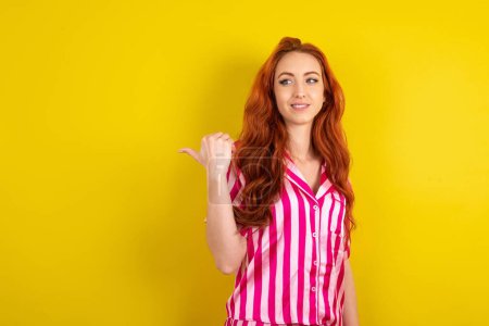 Téléchargez les photos : Jeune femme aux cheveux roux portant un pyjama rose sur fond jaune studio montrant à l'espace de copie, concept de publicité - en image libre de droit