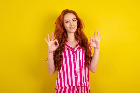 Foto de Mujer de pelo rojo joven con pijama rosa sobre fondo de estudio amarillo que muestra ambas manos con los dedos en signo OK. Aprobación o recomendación del concepto - Imagen libre de derechos