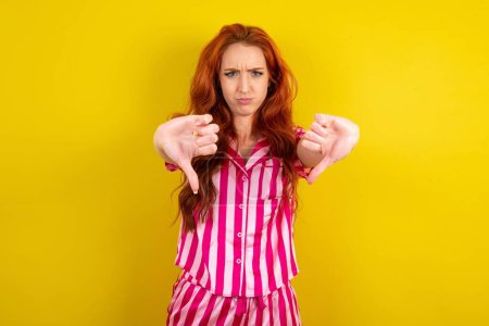 Téléchargez les photos : Jeune femme rousse portant un pyjama rose sur fond jaune studio étant bouleversé montrant pouce vers le bas avec deux mains. N'aime pas le concept. - en image libre de droit