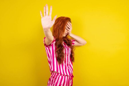 Téléchargez les photos : Jeune femme rousse portant un pyjama rose sur fond jaune studio couvre les yeux avec la paume et faire un geste d'arrêt, essaie de se cacher de tout le monde. - en image libre de droit