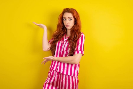 Téléchargez les photos : Jeune femme rousse portant un pyjama rose sur fond jaune studio pointant de côté avec les deux mains montrant quelque chose d'étrange et disant : Je ne sais pas ce que c'est. Concept de publicité. - en image libre de droit