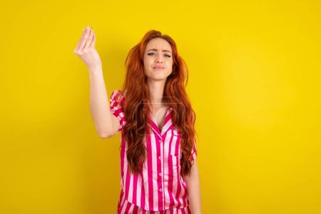 Téléchargez les photos : Jeune femme aux cheveux rouges portant un pyjama rose sur fond jaune studio geste de colère geste italien typique avec la main, regardant vers la caméra - en image libre de droit