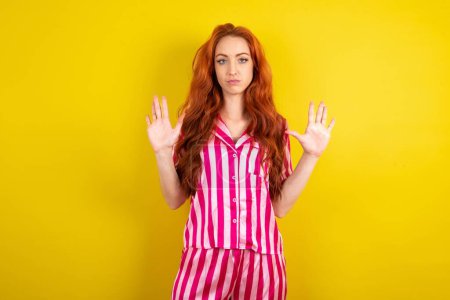 Photo for Young red haired serious woman wearing pink pyjama over yellow studio background pulls palms towards camera, makes stop gesture, asks to control your emotions and not be nervous - Royalty Free Image
