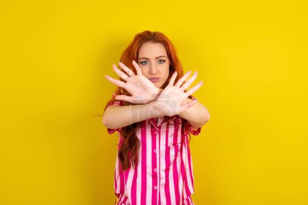 Téléchargez les photos : Jeune femme rousse portant un pyjama rose sur fond jaune studio a expression de rejet traversant les bras et les paumes faisant signe négatif, visage en colère. - en image libre de droit