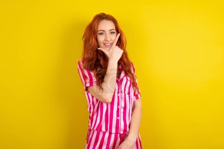 Téléchargez les photos : Jeune femme rousse portant un pyjama rose sur fond jaune studio regardant avec confiance la caméra souriant avec les bras croisés et la main levée sur le menton. Penser positif. - en image libre de droit