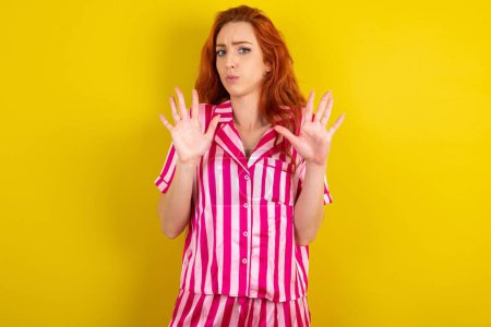 Téléchargez les photos : Jeune femme rousse portant un pyjama rose sur fond jaune studio éloignant les mains paumes montrant refus et déni avec une expression effrayante et dégoûtante. Arrêt et interdit. - en image libre de droit