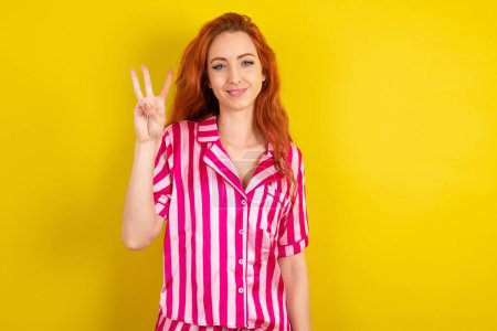 Téléchargez les photos : Jeune femme aux cheveux rouges portant un pyjama rose sur fond jaune studio montrant et pointant vers le haut avec les doigts numéro trois tout en souriant confiant et heureux. - en image libre de droit