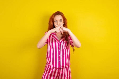 Téléchargez les photos : Jeune femme aux cheveux rouges portant un pyjama rose sur fond jaune studio a rejet expression en colère croisement des doigts faire signe négatif. - en image libre de droit
