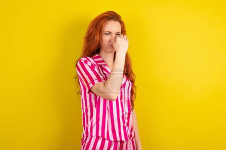 Téléchargez les photos : Jeune femme rousse portant un pyjama rose sur fond jaune studio sentant quelque chose de puant et dégoûtant, odeur intolérable, retenant son souffle avec les doigts sur le nez. Mauvaise odeur - en image libre de droit