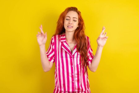 Téléchargez les photos : Jeune femme rousse portant un pyjama rose sur fond jaune studio geste doigt croisé souriant avec espoir et les yeux fermés. La chance et le concept superstitieux. - en image libre de droit