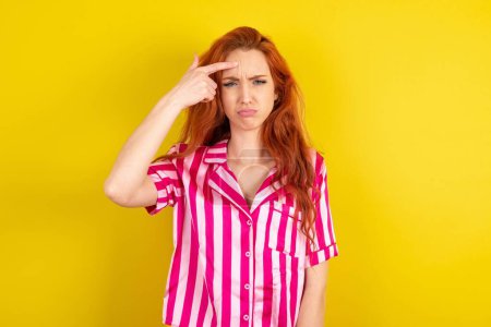 Téléchargez les photos : Jeune femme rousse portant un pyjama rose sur fond jaune studio pointant malheureux à bouton sur le front, infection du point noir. Concept de soins de la peau. - en image libre de droit