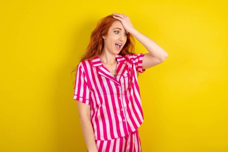 Téléchargez les photos : Jeune femme rousse portant un pyjama rose sur fond jaune studio surpris avec la main sur la tête pour erreur, rappelez-vous erreur. Oublié, mauvais concept de mémoire. - en image libre de droit