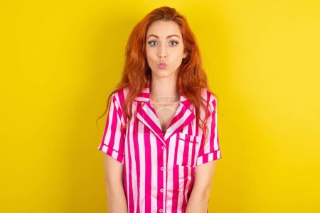 Photo for Shot of pleasant looking red haired woman wearing pink pyjama over yellow studio background, pouts lips, looks at camera, Human facial expressions - Royalty Free Image