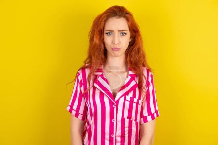 Téléchargez les photos : Mécontent bouleversé femme aux cheveux rouges portant pyjama rose sur fond jaune studio fronce les sourcils visage comme va pleurer, être mécontent et malheureux comme ne peut pas atteindre les objectifs, modèle déçu a des problèmes - en image libre de droit