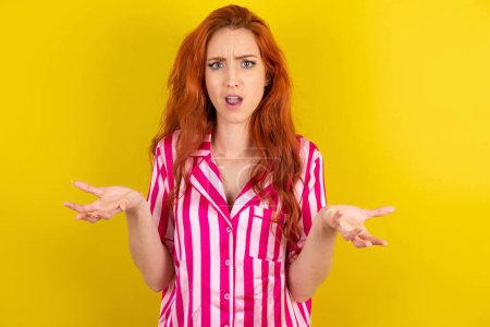Photo for Frustrated red haired woman wearing pink pyjama over yellow studio background feels puzzled and hesitant, shrugs shoulders in bewilderment, keeps mouth widely opened, doesn't know what to do. - Royalty Free Image