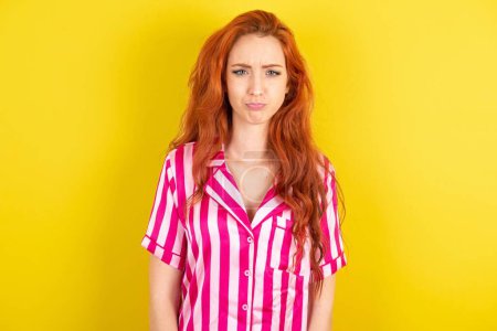 Photo for Dismal gloomy rejected red haired woman wearing pink pyjama over yellow studio background has problems and difficulties, curves lower lip and closes eyes in despair, being in depression - Royalty Free Image