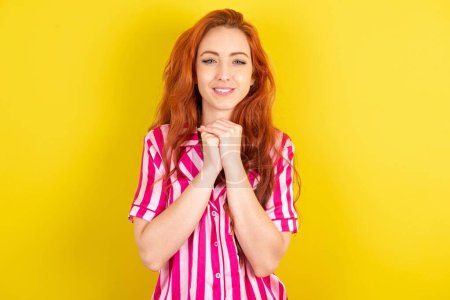 Photo for Positive red haired woman wearing pink pyjama over yellow studio background smiles happily, glad to receive pleasant news from interlocutor, keeps hands together. People emotions concept. - Royalty Free Image