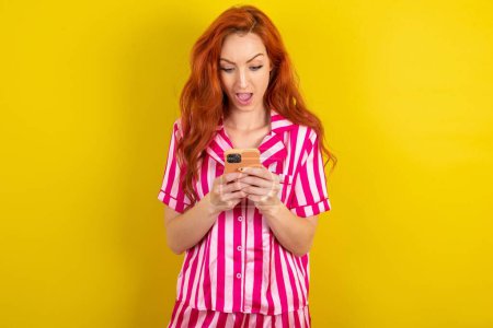 Téléchargez les photos : Femme aux cheveux rouges portant un pyjama rose sur fond jaune studio en utilisant le téléphone mobile bavarder temps libre . - en image libre de droit
