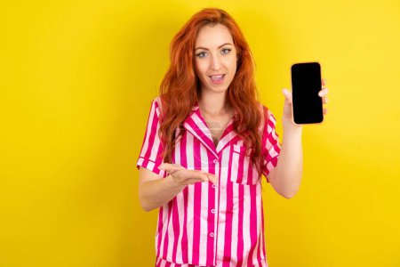 Photo for Young red haired woman wearing pink pyjama over yellow studio background with a mobile presenting smartphone. Advertisement concept. - Royalty Free Image