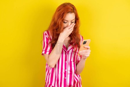 Photo for Young red haired woman wearing pink pyjama over yellow studio background looking at smartphone feeling sad holding hand on face. - Royalty Free Image