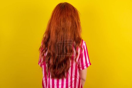 Foto de Joven mujer pelirroja con pijama rosa sobre fondo de estudio amarillo que muestra la vista trasera. Studio Shoot. - Imagen libre de derechos