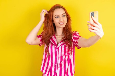 Foto de Joven mujer pelirroja vistiendo pijama rosa sobre fondo de estudio amarillo sonriendo y tomando una selfie lista para publicarla en sus redes sociales. - Imagen libre de derechos