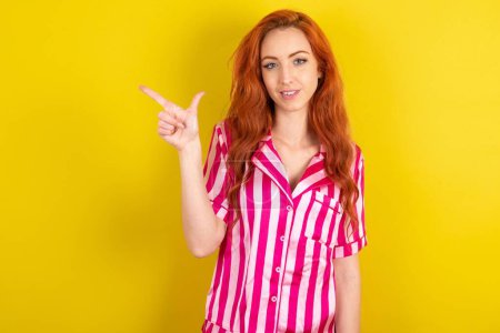 Photo for Young red haired woman wearing pink pyjama over yellow studio background pointing up with fingers number eight in Chinese sign language B. - Royalty Free Image
