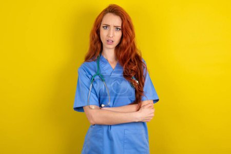 Téléchargez les photos : Choqué embarrassé jeune femme médecin aux cheveux roux sur fond jaune studio garde la bouche largement ouverte. - en image libre de droit