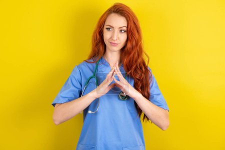 Photo for Young red-haired doctor woman over yellow studio background steepled fingers and looks mysterious aside has great evil plan in mind - Royalty Free Image