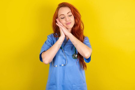 Photo for Young red-haired doctor woman over yellow studio background leans on pressed palms closes eyes and has pleasant smile dreams about something - Royalty Free Image